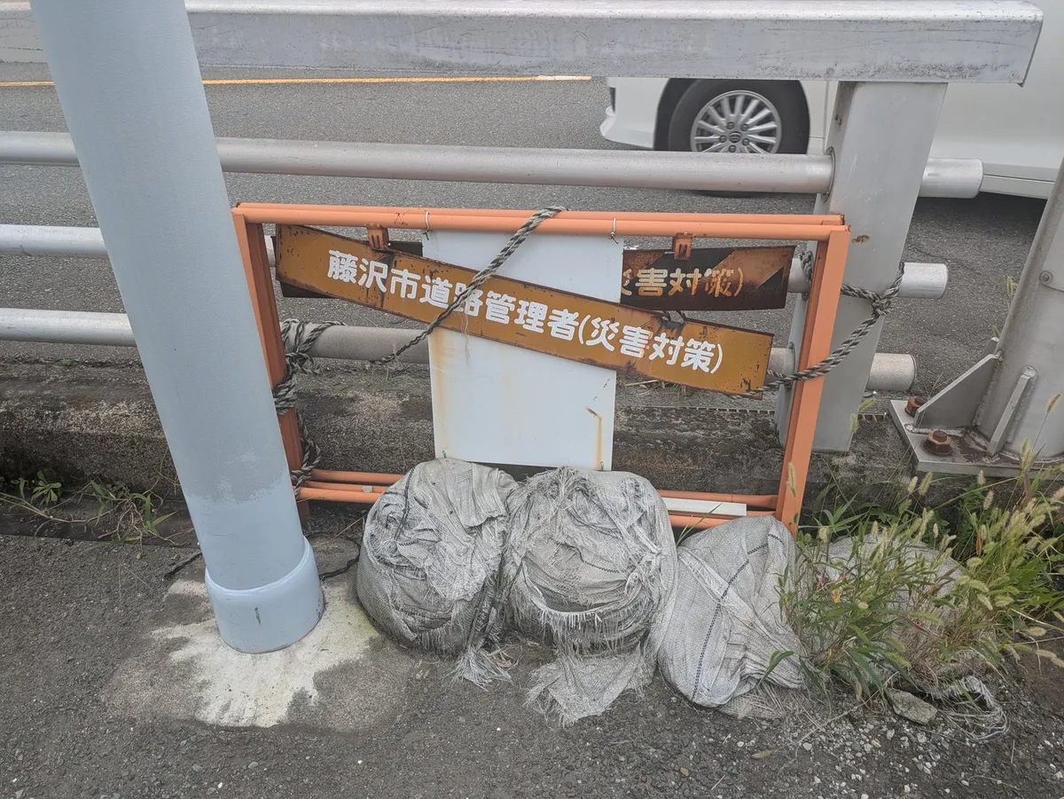 道路脇の柵に「藤沢市道路管理者（災害対策）」の板が付き、足元に土のうが3つ置かれている